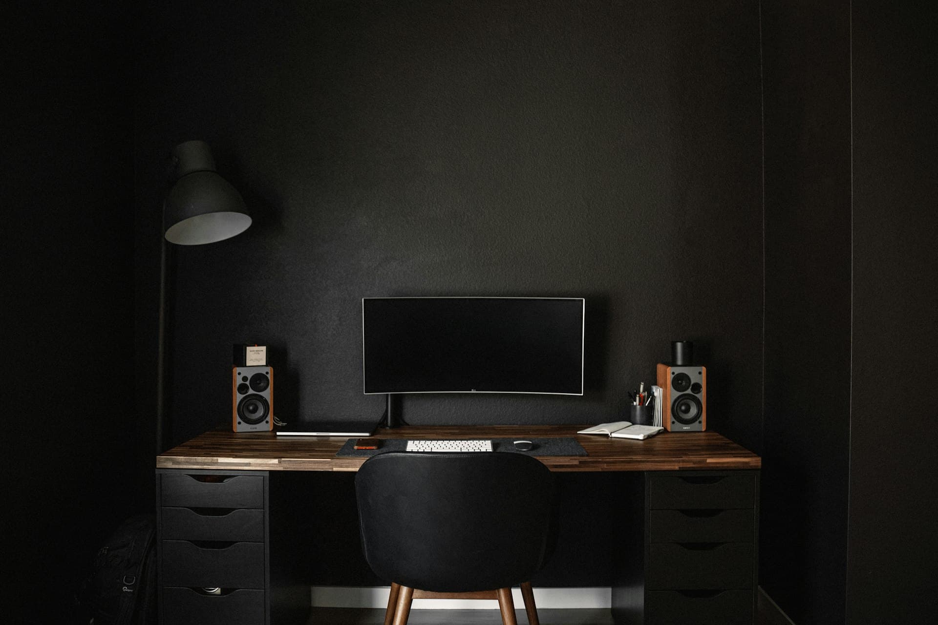 A desk against a black wall with an ultrawide monitor, two speakers, an open notebook and a floor lamp.